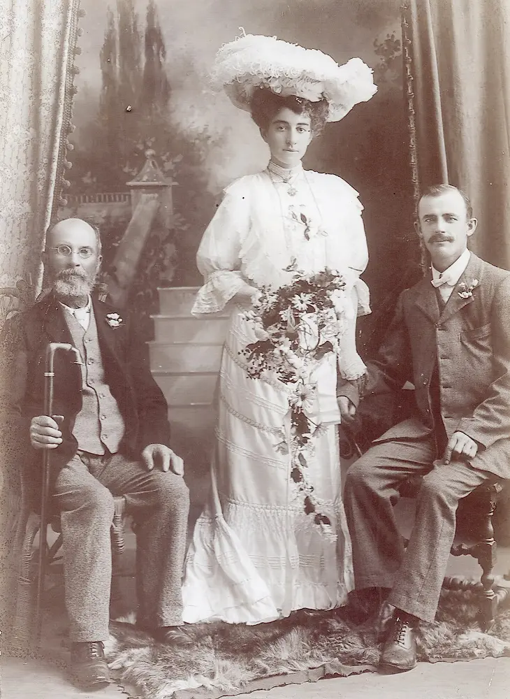 An old, black and white photo portrait. An older gentleman sits on the left, a young woman stands in the centre, and a young man sits on the right.