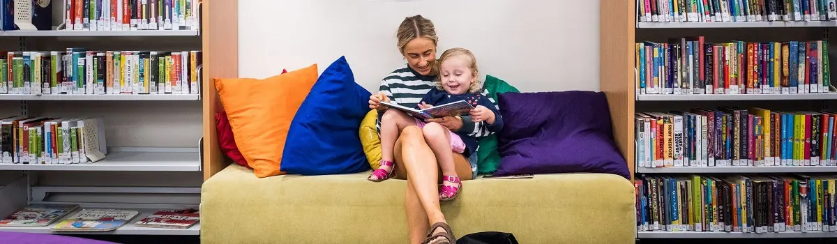 A woman in the library sitting down and reading a book to a small child.